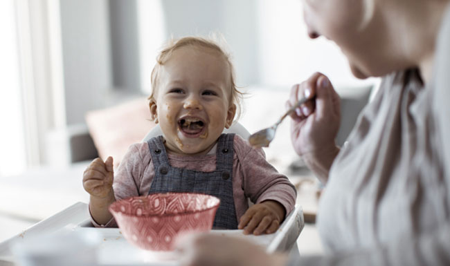caregiver feeding happy young infant at the table with a spoon